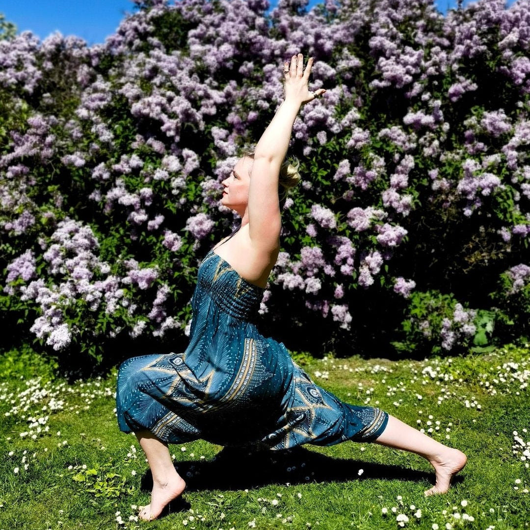 A woman in a blue patterned strapless dress performs a yoga pose on a grassy lawn with a large purple flowering bush and blue sky behind her.